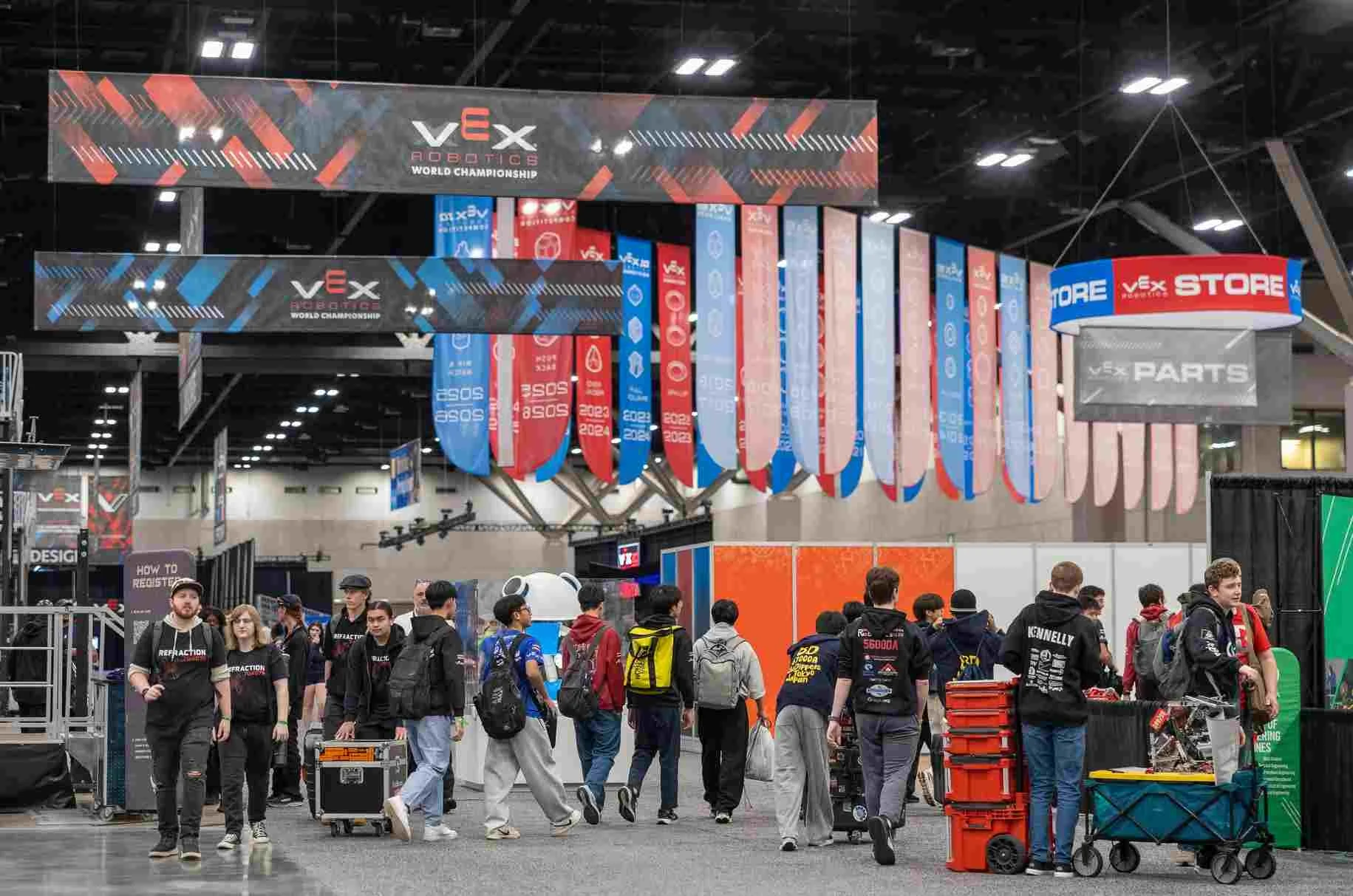 Teams moving robots and kit crates across the pit floor under the VEX Robotics banners