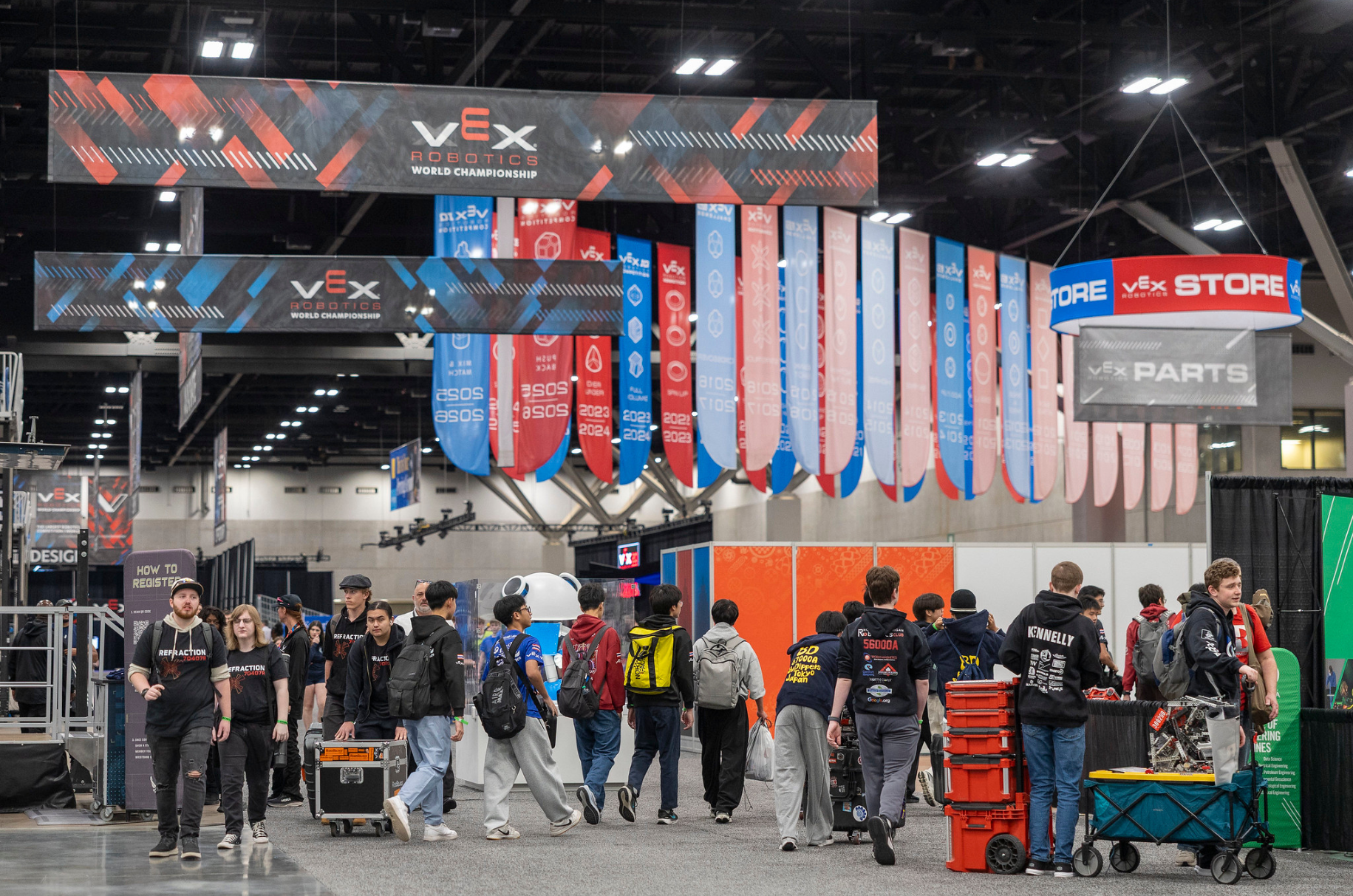 Teams moving robots and kit crates across the pit floor under the VEX Robotics banners