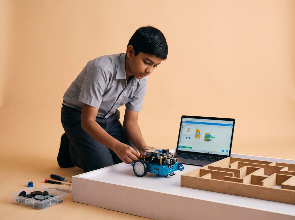 A student adjusting a wheeled robot beside a maze, with block code open on a laptop