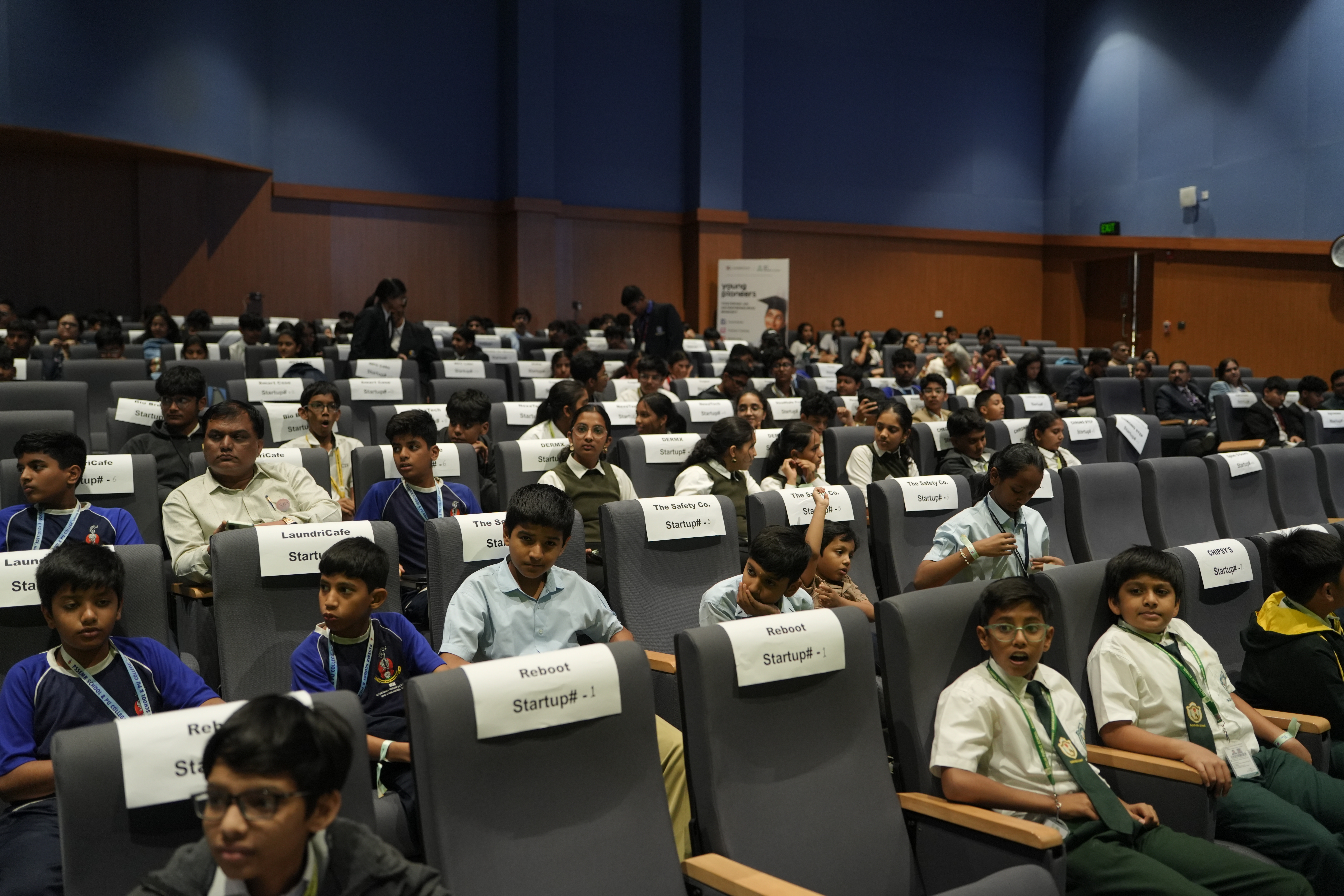 Teams seated in the auditorium, each seat labelled with a startup name and number