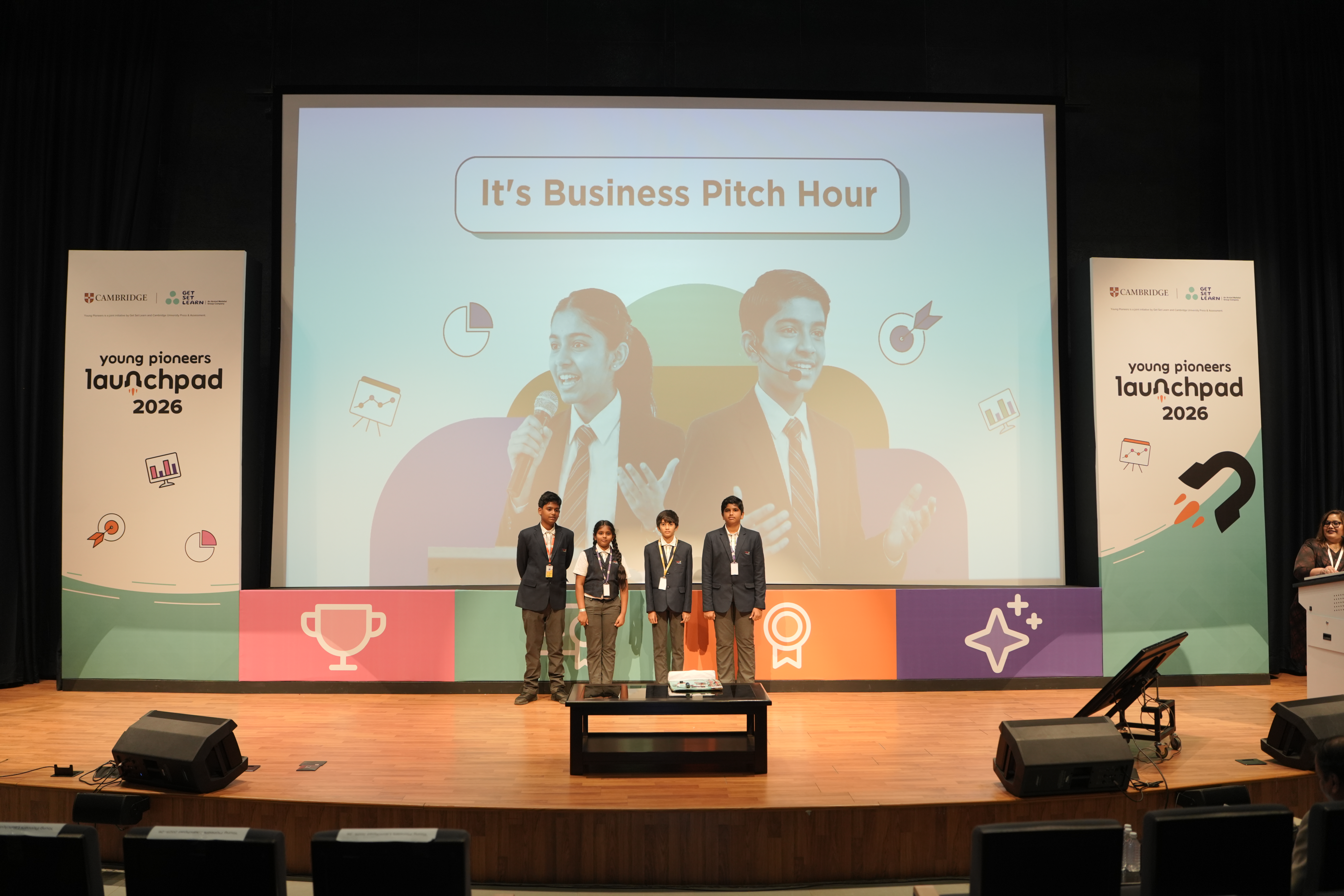 A team of four students on stage at Launchpad 2026, with the business pitch hour slide behind them
